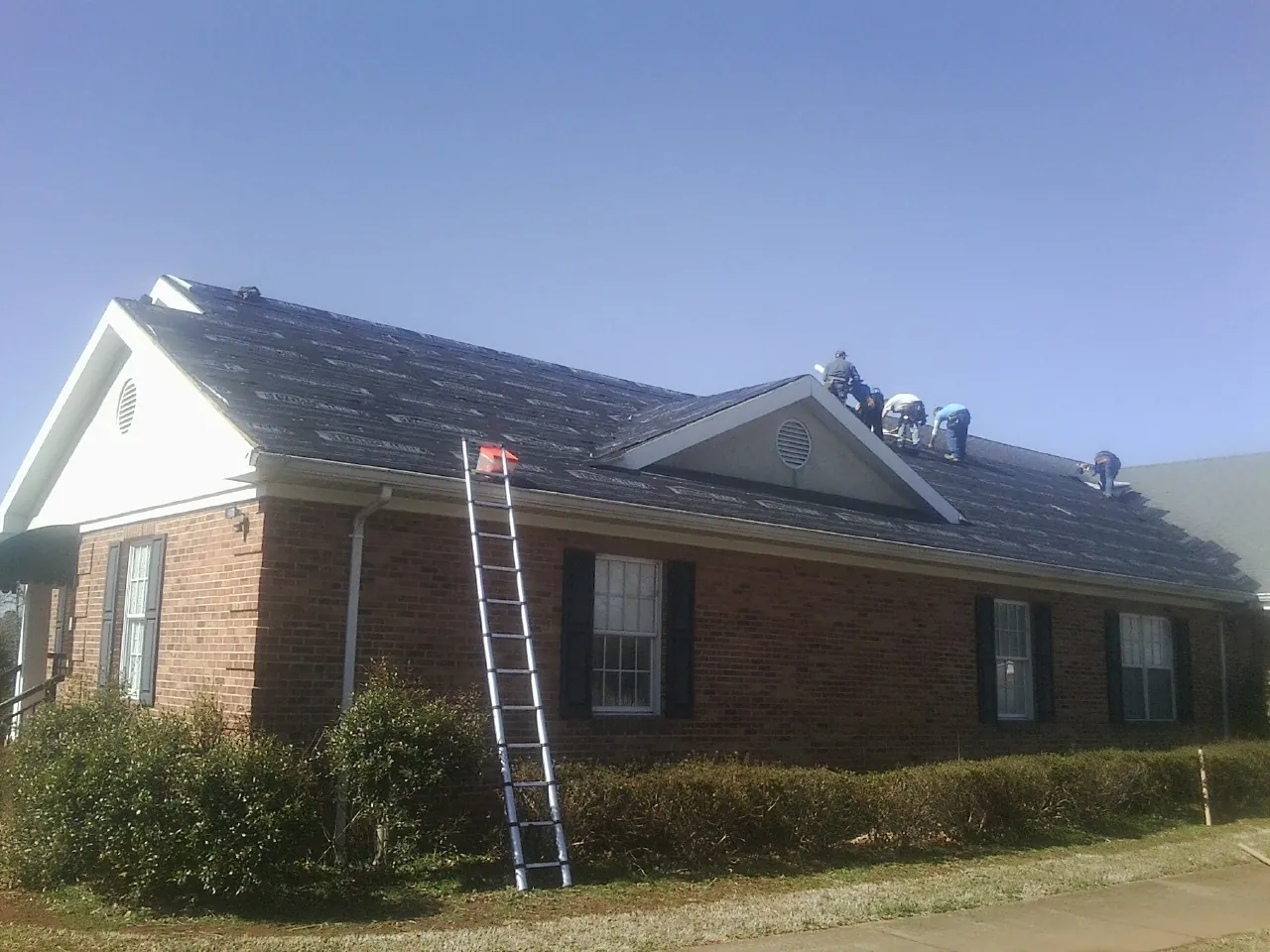 Crew tearing shingles off a brick ranch roof with a ladder set — Emergency Roof Repair work by our crew in Travelers Rest