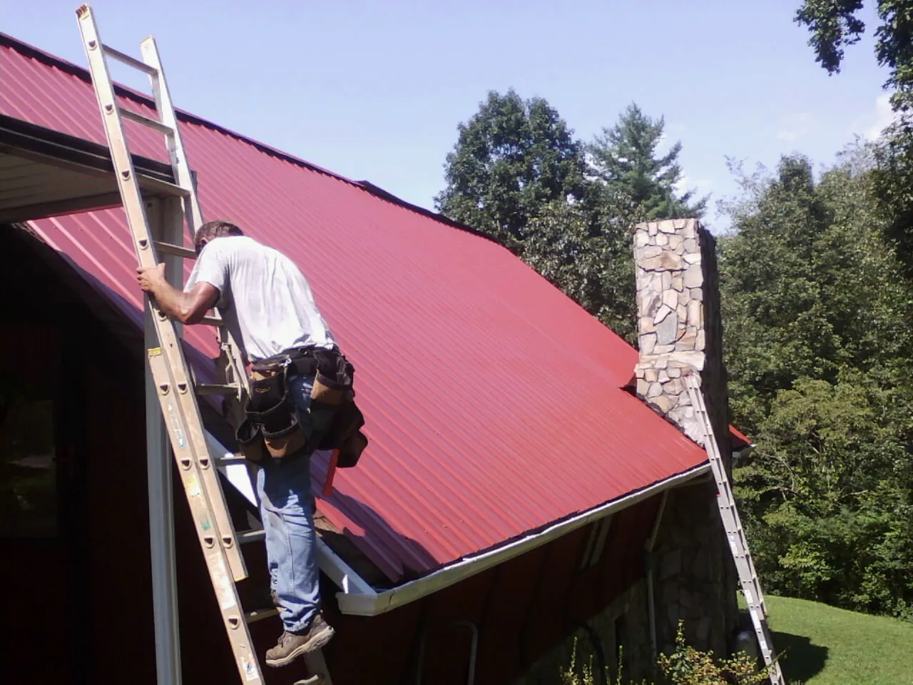 Roofer on a ladder beside a red metal roof and stone chimney — Soffit & Fascia Repair by Roofing Brothers in Travelers Rest